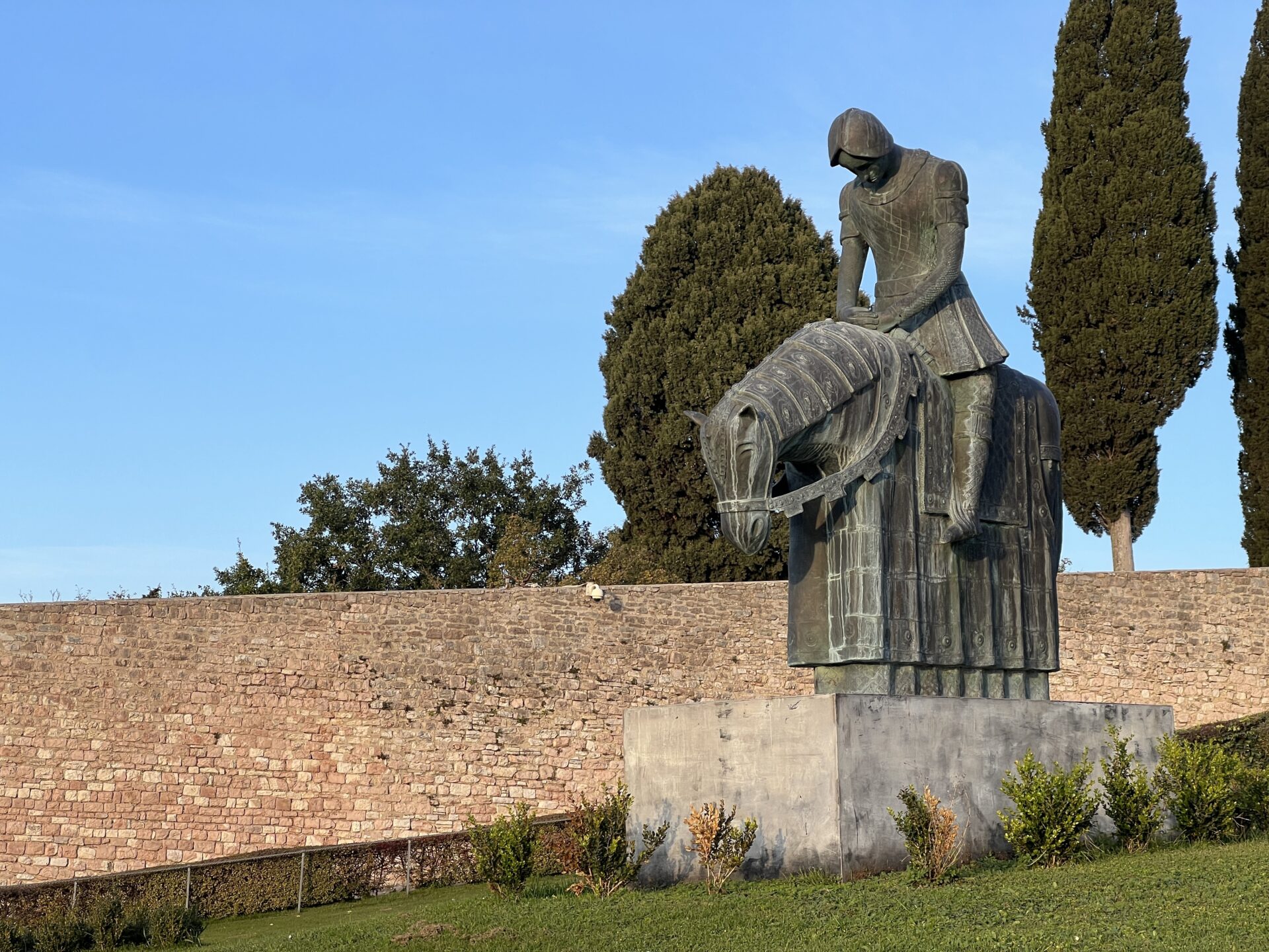 Statue of knight on horseback near stone wall and trees.